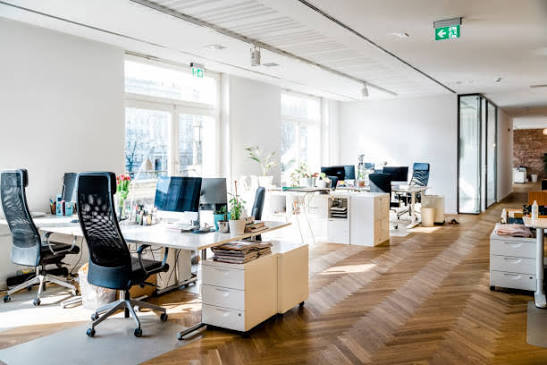 A bright, modern open-plan office space with white desks, black mesh office chairs, computer monitors, and a herringbone wood floor. Large windows on the left let in natural daylight, and a green exit sign is visible on the ceiling.